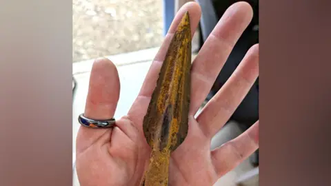 Headland Archaeology A medieval spearhead made of iron, held in a hand, which bears a ring on the thumb. The spear head has four fins, three of which are visible. It is showing signs of rusting, but is well preserved, and shaped to a point at the tip and curved towards the back end.