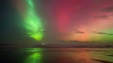Green, pink, orange and pale yellow light glows above the beach and is reflected in the sea and wet sand.
