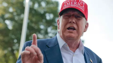 Donald Trump wearing a white shirt and navy suit jacket with no tie. He is wearing a red cap which bears the words: GULF OF AMERICA. Yet another TRUMP development.