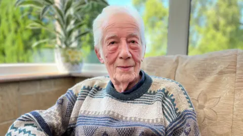 90-year-old Ernest Williams sat in a conservatory. There are large windows behind him showing blue sky and trees outside. 