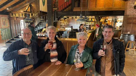 BBC Four people holding glasses are by a table smiling and looking at the camera. A bar and stairs are in the background.