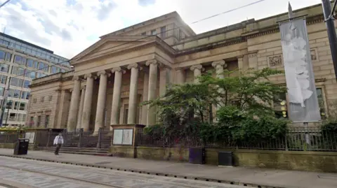 Google A street view of a large sand coloured stone building. The building has columns and is build in an ancient Greek/Roman architectural style.