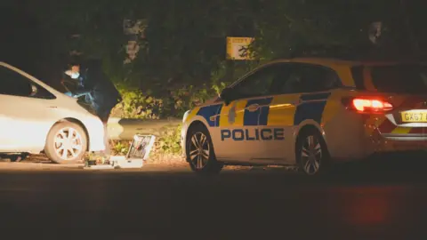 Eddie Mitchell A police officer investigating a white car. It is dark and they are wearing black clothing and blue latex gloves. A police car is parked next to the scene.
