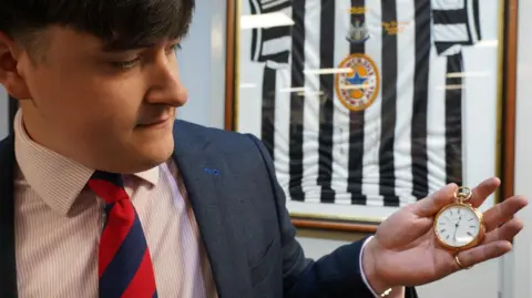 Jack Orgill holding a gold pocket watch in his outstretched hand. He has a gold thin ring on his pinkie finger. He is wearing a blue suit with a pink and white striped shirt and blue and red diagonally striped tie. He has brown hair which reaches his eyebrows. There is a 1997 black and white Newcastle United shirt hanging in a frame in the background.