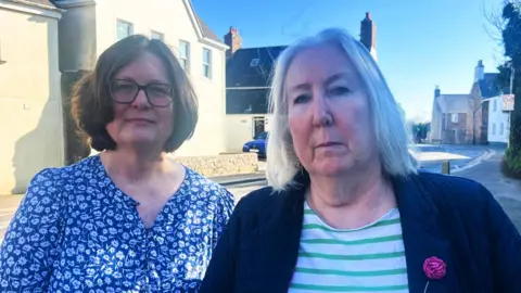 Alison Marshall and Jane Reed are middle-aged white women. They are standing by a road with houses behind them. They are both looking into the camera. Alison is wearing a blue and white blouse, Jane is wearing a green and white striped top with a blue jacket on top.