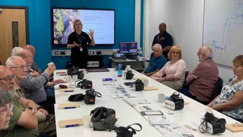 Men and women sit around a table with notebooks and virtual reality headsets in front of them. A woman is talking to them in front of a screen.