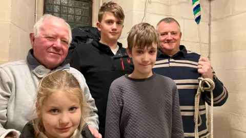 Five people stood in an indoor setting smiling at the camera. On holds a rope leading up to the ceiling. There are three children and two adults