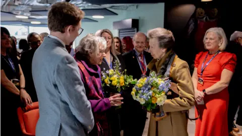 University of Gloucestershire Edna Lee speaks with Princess Anne. Both hold bunches of flowers and are surrounded by university staff. The princess stands on the right and wears a brown coat and Edna wears a purple one