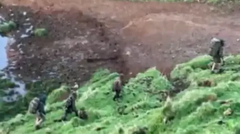 NZ police Phillips and his three children, in this blurry image, seen wearing camouflage uniforms and carrying large backpacks, trek through green bush with bare earth and some water visible
