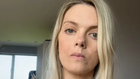 A selfie of Elen Hughes inside a house with light grey walls and a window visible behind her. She looks serious and has long blonde hair and there is a light grey cupboard behind her