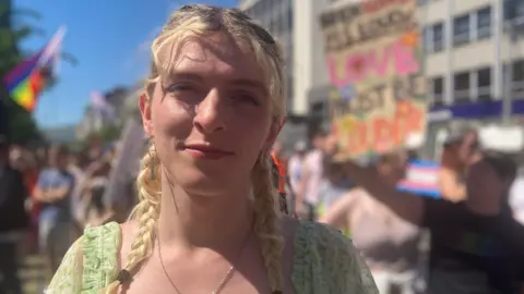 Alexa is wearing a green dress and silver necklace.
Their hair is blond in two plaits with dark hair coming through the roots.
Behind is a blurred out rally.