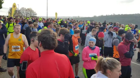 Oli Constable/BBC A large group of runners in a variety of brightly colored vests, sports tops and leggings huddle together at the start line.