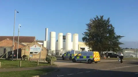 Two police vans are parked outside a factory site with brick buildings and tall, white containers seen behind metal fences under bright blue skies, with a white sign on grass in the foreground saying Banham Poultry Limited.