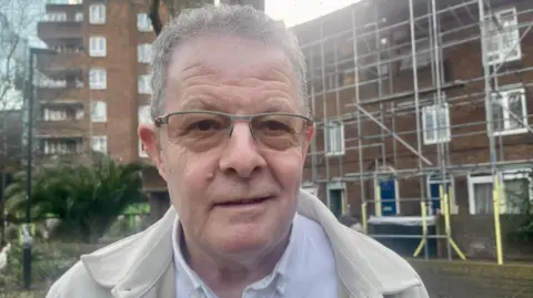 A white man with grey hair and a cream jacket stands on a council estate which is covered in scaffolding
