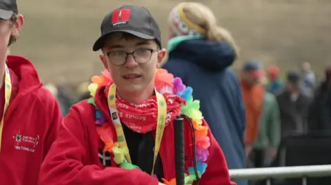 Alfie smiling as he has his photo taken. He is wearing a red jumper, with a Hawaiian flower necklace. He is wearing a black baseball cap and glasses. There are crowds of people stood behind him. 