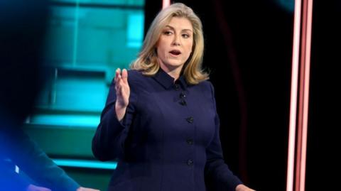 Penny Mordaunt in a navy blue suit, looking to her left with arm and hand extended. She has shoulder-length blonde hair