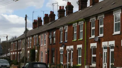 Christopher Hilton/Geography Crown Street in Stowmarket, showing a row of brick built terrace houses, all with windows and front doors. There is a road in front of the houses with two cars parked on the road. There are two telegraph poles and chimneys on the roofs. 