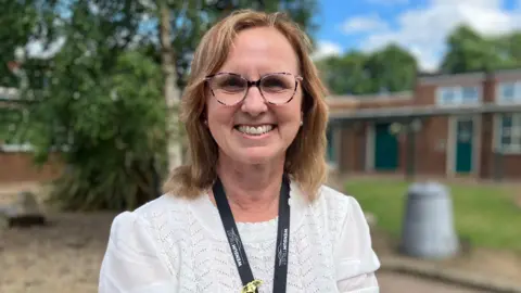 Owen Sennitt/BBC Rosalind Robinson, the executive head teacher at Firside Junior School. She is standing outside in front of the school and is looking directly at the camera and smiling. She is wearing a white patterned blouse with a black school lanyard round her neck and a pair of glasses on her eyes.