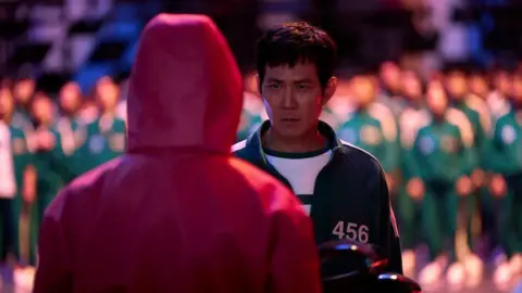 No Ju-han/Netflix Lee Jung-jae as Gi-hun, wearing a green tracksuit and frowning at a guard wearing a hooded red suit.