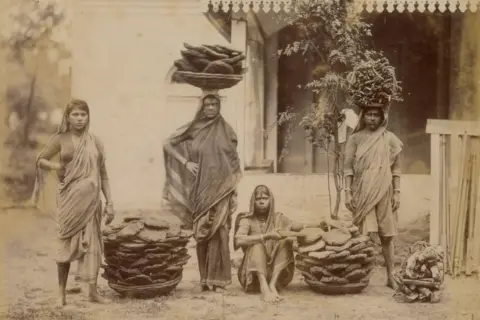 DAG Five Indian women wearing saris stand outside a modest house in Bombay (now Mumbai) in 1890, balancing neatly stacked cow dung cakes on their heads while additional rounds lie arranged on the ground beside them.

