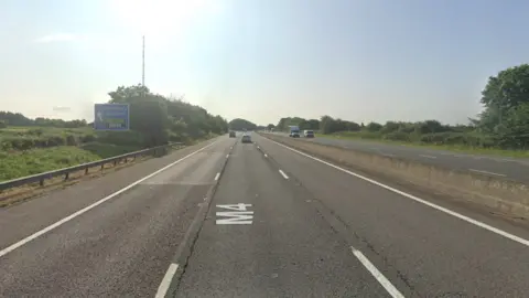 Google maps street view of the M4 eastbound carriageway near the exit for Membury Services. There is a blue sign pointing left for the services, with a black, green and white Welcome Break logo underneath. The road has three lanes and a hard shoulder. A few vehicles can be seen travelling eastbound and there are some on the westbound carriageway too. Trees and fields can be seen either side of the motorway. 