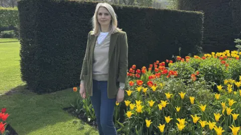 Lady Antrim wearing a green jacket, cream jumper and white top. She has blue jeans and has blonde hair. She is surrounded by tulips that are yellow and orange and shrubbery