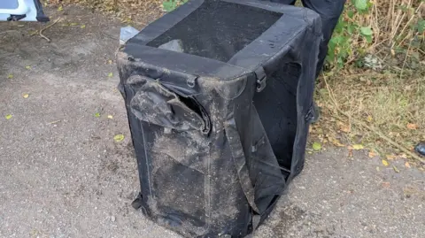 A black container with netted sides which is on the ground. It is wet and covered in sand and dirt.