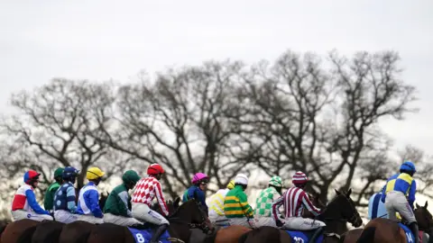 PA Media Jockeys are sitting on horses in a row. They are seen from the back, all of them are wearing colourful jerseys.