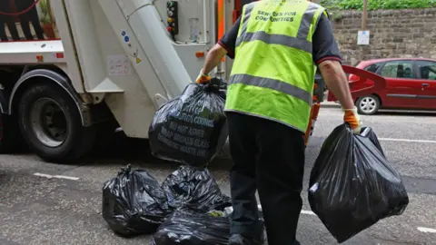 A person wearing a yellow high-vis vest carries bin bags in either hands towards the rear of a bin lorry parked on a road. His body and legs are in shot but his head is not. There are a small pile of bin bags at his feet.