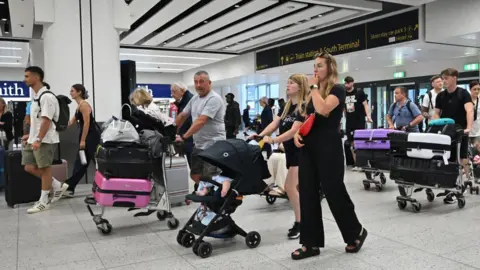 Getty Images A busy airport concourse with a woman pushing a pushchair in the foreground and various other passengers pushing trolleys and cases. 