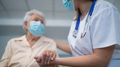 A medical professional holds the hand of an elderly person. Both are wearing face masks. The elderly person is wearing a light peach-coloured shirt and the medical professional is wearing a white top and has a stethoscope around their neck.