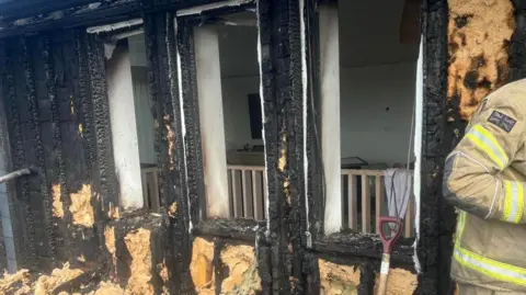 Cornwall Fire Service Charred exterior walls which have been turned black following a fire in Cornwall. Part of a firefighter in uniform can be seen on the right-hand side of the photo and the top half of a shovel is propped against one of the walls.