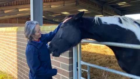 Claire Dickie is petting a horse's head, which is looking over a stable gate. Ms Dickie is wearing a blue jacket. The horse is dark brown with white markings.