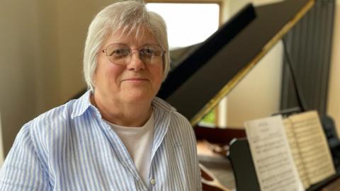 Ann is a white woman with short, grey hair and she is wearing glasses. She is stood next to her late husband's grand piano.