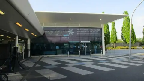 GEOGRAPH/JONATHAN THANKER Single-storey modern railway station building, with glass frontage and a white portico. There are advertising slogans on the building. There is a black and white paved area in front of the station and some fir trees to the right.