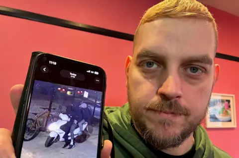A man with short ginger hair stands in a room in front of a red-coloured wall while holding his phone up to the camera. An image on the screen of the phone shows three youths wearing balaclavas sitting on the back of a white moped at night. The man is wearing a green hoody and has a serious expression. 
