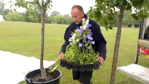 A man in a dark top walks between two trees carrying pots of flowers.