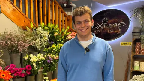 Jamie is wearing a blue quarter zip sweater. He is smiling and behind him is an array of flowers and plants.