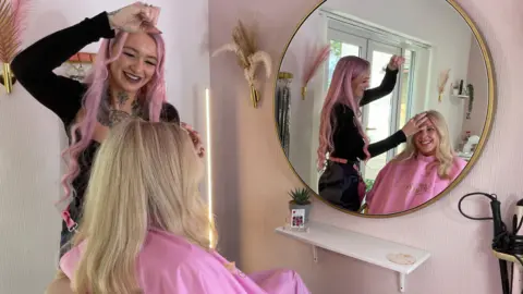 Taylor Keig has long wavy pink hair and a large tattoo on her neck and chest. She is smiling and looking down at Laura Rush in the hair salon. Taylor is doing Laura's hair and you can see Laura smiling, wearing a pink robe, in the mirror reflection in front of her.