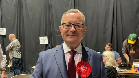 BBC Lee Barron with short white hair wearing a red rosette and tie and a grey jacket and standing in front of a black curtain at an election count