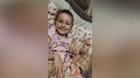 Family A young girl with short brown hair and a medical tube taped to her face grins at the camera. She lies down with a small dog cuddled into her arm.