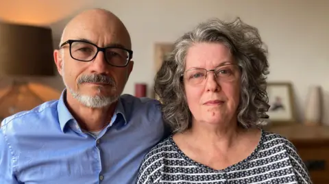 Paul and Andrea face the camera straight on. Paul is bald with a grey goatee and short beard and wears glasses, he is wearing a light blue shirt. Andrew has wavy grey hair and glasses and is wearing a patterned white and black top. A dresser with pictures on can be seen blurred in the background. 