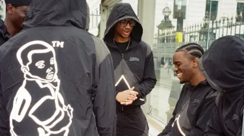 Five young men in black tracksuits stand at a bus stop