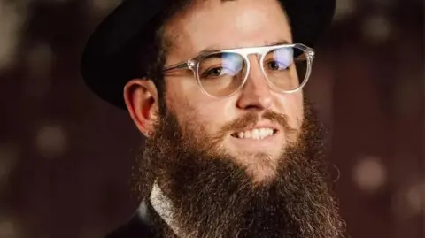 Zvi Kogan is seen in a portrait photograph. A bearded man, he is smiling at the camera and wearing glasses with clear plastic frames. There is a black hat, a fedora, on his head.