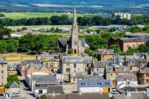 Getty Images A general view of Invergordon. There are a variety of one and two-storey buildings in the foreground, and a church with a large steeple in the middle of the picture. Beyond that is an industrial estate surrounded by green fields and trees.