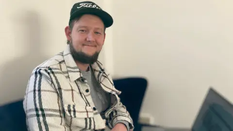 Mr Digby, digital inclusion manager, is facing the camera and smiling. He is a white man with brown hair and a brown beard. He is wearing a black cap, a grey T-shirt, and a cream jacket with black and brown tartan stripes.