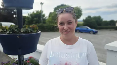 A woman in a white t-shirt is standing beside a flower pot, she has sunglasses on her head. behind her there is a car park with one blue car parked in it.