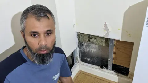 A man wearing a navy blue and light blue colour T-shirt, with small white writing in the top left. He is positioned near two square cut holes inside the interior of his living-room walls. Inside these holes you can see wood and other materials. The living-room walls are white and the flooring is wooden. 