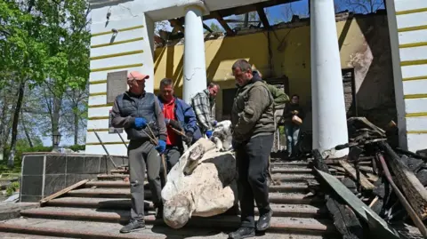 SERGEY BOBOK/AFP Pekerja museum membawa patung filsuf Ukraina Hryhorri Skovoroda dari bangunan Museum Peringatan Sastra Nasional Hryhoriy Skovoroda yang hancur di desa Skovorodynivka, di Wilayah Kharkiv, pada 7 Mei 2022