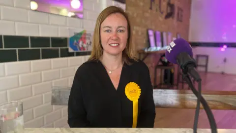 A woman with mid-length brown-blonde hair sits at a table with purple lighting in the background. She is wearing a black shirt and has a yellow rosette pinned to her top.